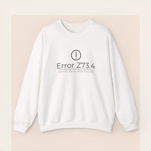 May include: White crew neck sweatshirt featuring the text "Error Z73.4 Social Skills Not Found" in black, with an exclamation mark above. A casual, unisex garment, perfect for everyday wear.