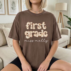 以下が含まれることがあります： フローラルデザインの「First Grade」の文字と、筆記体で「miss molly」と書かれた茶色のTシャツ。半袖のカジュアルスタイルです。