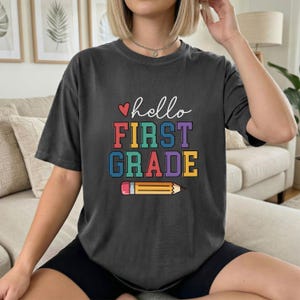 以下が含まれることがあります： 「hello FIRST GRADE」の文字と鉛筆のイラストがプリントされた濃いグレーのTシャツ。カジュアルなスタイルで、新学期や先生へのギフトに最適です。