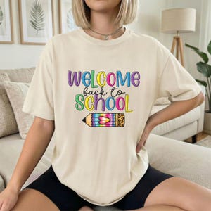 以下が含まれることがあります： カラフルな文字で「WELCOME back to SCHOOL」と書かれたベージュ色のTシャツ。テキストの下には虹色の鉛筆のグラフィックがあります。 ゆったりとしたフィット感のシャツです。