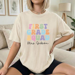 以下が含まれることがあります： ベージュ色のTシャツ。カラフルなバブル文字で「FIRST GRADE SQUAD」と黒い筆記体で「Mrs. Gibson」と書かれています。カジュアルでオーバーサイズのスタイルです。