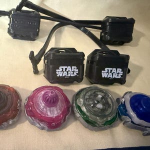 May include: Four spinning tops in red, purple, green, and blue with metallic silver bases. Above them are two black plastic launchers with Star Wars logos and two black pull-string cords. The items are arranged on a light-colored fabric surface.