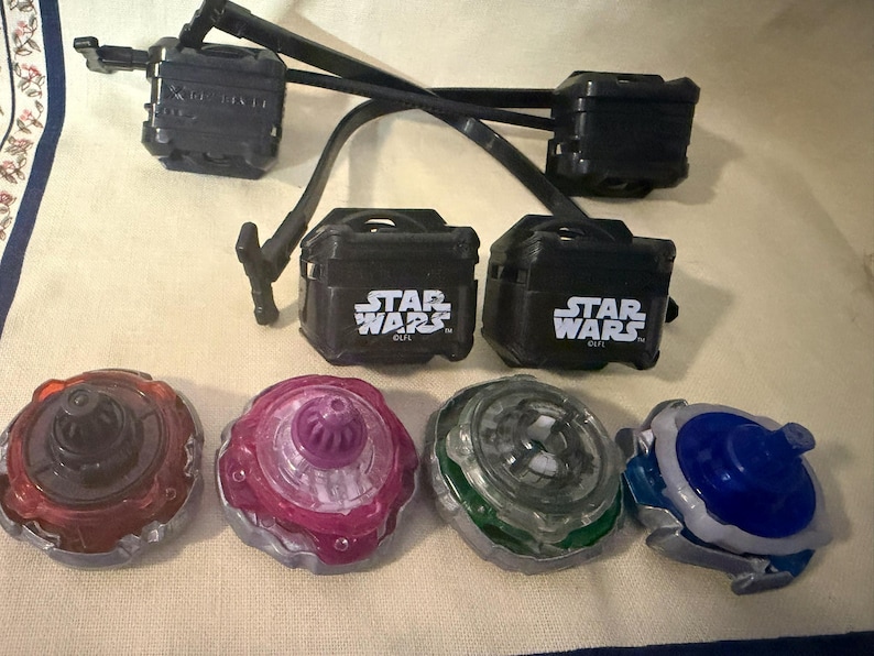 May include: Four spinning tops in red, purple, green, and blue with metallic silver bases. Above them are two black plastic launchers with Star Wars logos and two black pull-string cords. The items are arranged on a light-colored fabric surface.