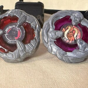 May include: Two Beyblade spinning tops featuring metallic silver outer rings. The left top has a red translucent center with a Darth Vader graphic. The right top has a purple translucent center with an orange and white creature graphic.