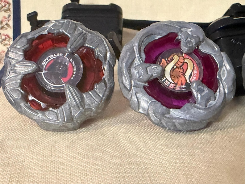May include: Two Beyblade spinning tops featuring metallic silver outer rings. The left top has a red translucent center with a Darth Vader graphic. The right top has a purple translucent center with an orange and white creature graphic.