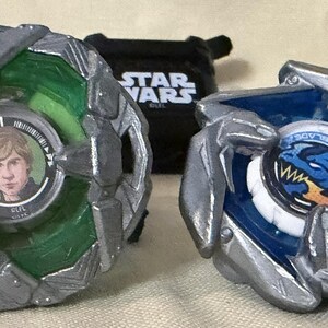 May include: Two Beyblade X spinning tops with silver metallic outer rings. The left top features a green translucent body with a circular center image of a person. The right top has a blue translucent body with a blue dragon graphic center.