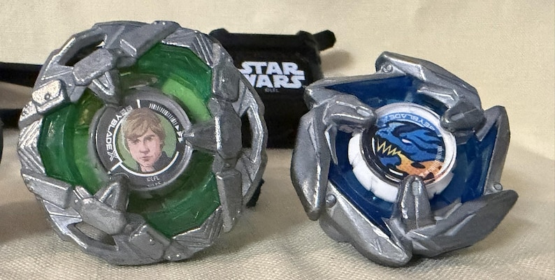 May include: Two Beyblade X spinning tops with silver metallic outer rings. The left top features a green translucent body with a circular center image of a person. The right top has a blue translucent body with a blue dragon graphic center.