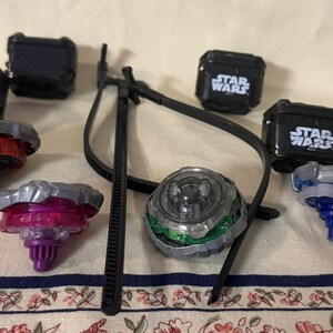 May include: A collection of four spinning tops with metallic and plastic components in red, purple, green, and blue, accompanied by four black square cases labeled Star Wars and a black plastic tool with a long, thin, curved handle and arm.