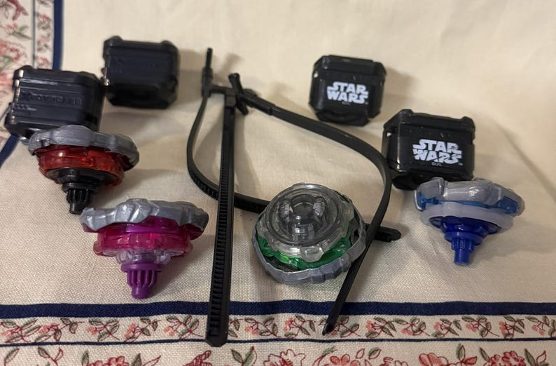 May include: A collection of four spinning tops with metallic and plastic components in red, purple, green, and blue, accompanied by four black square cases labeled Star Wars and a black plastic tool with a long, thin, curved handle and arm.