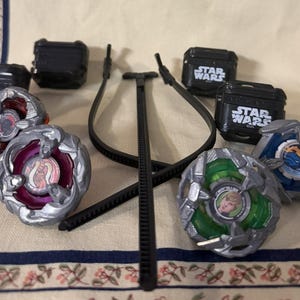 May include: A collection of four spinning top toys with metallic silver frames and colorful centers, including one with a green base featuring a character portrait. Four black plastic cases labeled Star Wars and a black plastic ripcord launcher are included.