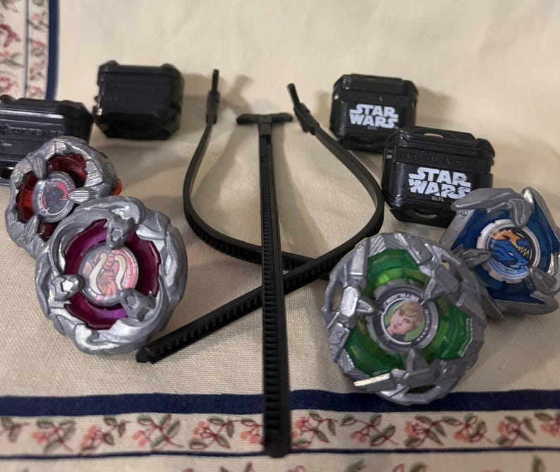 May include: A collection of four spinning top toys with metallic silver frames and colorful centers, including one with a green base featuring a character portrait. Four black plastic cases labeled Star Wars and a black plastic ripcord launcher are included.