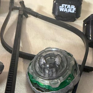 May include: A circular spinning top toy with a clear plastic center, a green inner ring, and a metallic outer rim. Nearby is a black plastic block with the white text STAR WARS and a long black plastic zip tie component with a T-shaped connector.