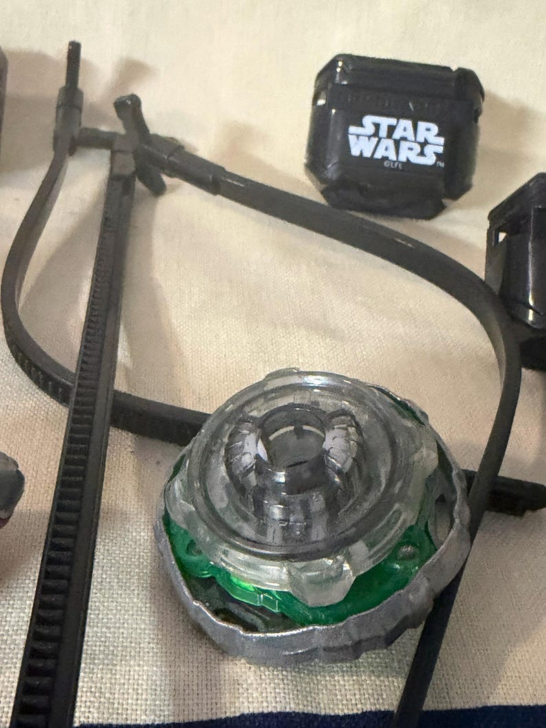 May include: A circular spinning top toy with a clear plastic center, a green inner ring, and a metallic outer rim. Nearby is a black plastic block with the white text STAR WARS and a long black plastic zip tie component with a T-shaped connector.