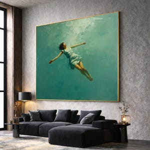 Op de afbeelding: Groot ingelijst kunstwerk met een vrouw in een lichtblauwe jurk die in turquoise water zweeft. Het schilderij heeft een gouden lijst en hangt boven een donkergrijze hoekbank. De kamer heeft een modern, minimalistisch design.