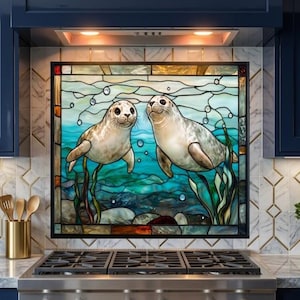 May include: A square stained glass panel featuring two light brown seals swimming underwater among green seaweed and bubbles. The artwork is framed in black and displays a detailed aquatic scene with blue and teal tones, positioned above a stainless steel stove.