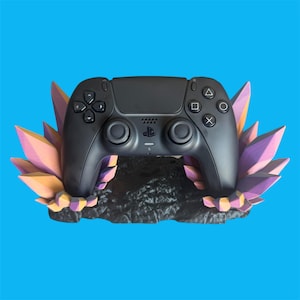 Peut inclure: Une manette noire PlayStation 5 posée sur un support décoratif. Le support présente une base sombre avec un design en cristal dans les tons rose, orange et jaune. La manette a une disposition symétrique des boutons.