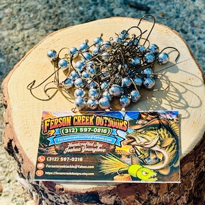 May include: A pile of silver fishing jig heads with bronze hooks rests on a wooden slice. A business card for Ferson Creek Outdoors is visible, featuring a fish illustration and contact information.