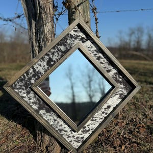 May include: Diamond-shaped mirror with a rustic wooden frame. The frame features a cowhide pattern in black and white. The mirror is leaning against a weathered wooden post, with a clear blue sky in the background.