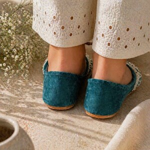 May include: Teal suede slip-on shoes with a beaded upper trim and a rounded toe. The shoes have a brown sole and are worn with cream-coloured trousers with eyelet detailing.