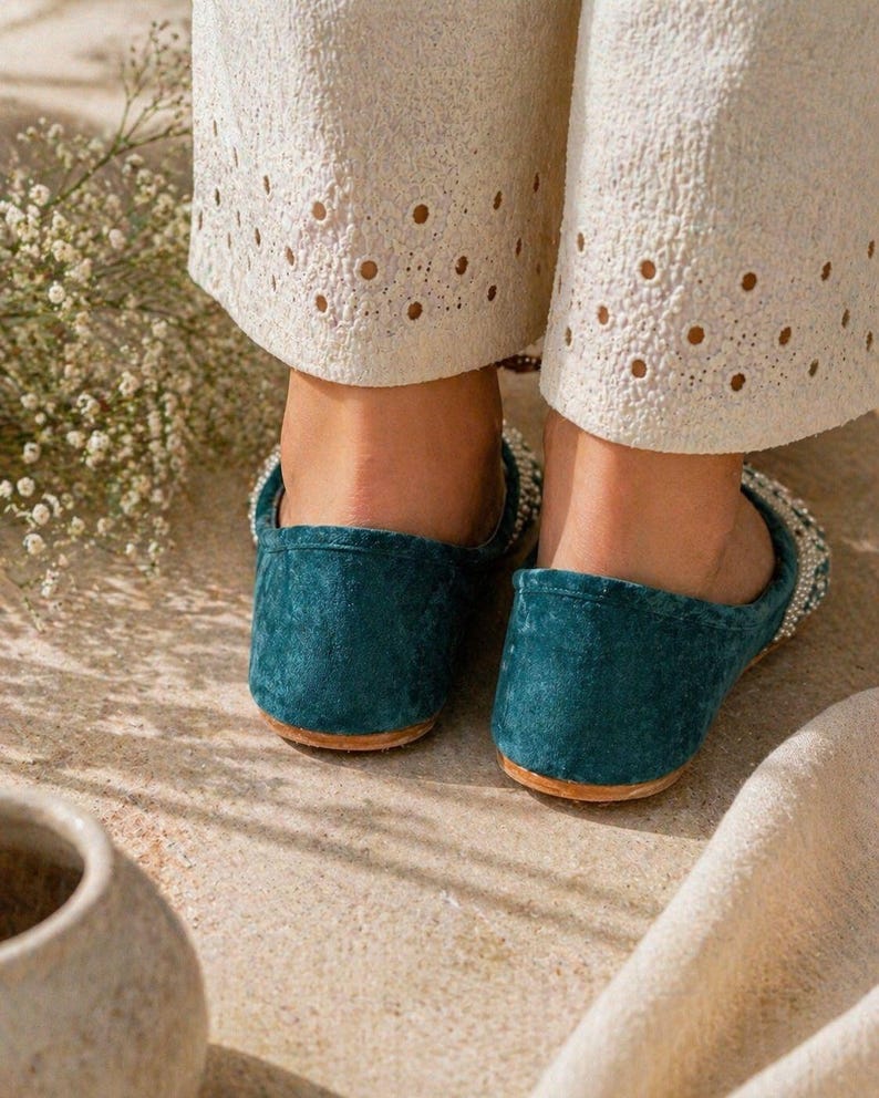 May include: Teal suede slip-on shoes with a beaded upper trim and a rounded toe. The shoes have a brown sole and are worn with cream-coloured trousers with eyelet detailing.