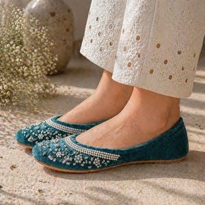 May include: Teal velvet flats adorned with pearls and beads. These shoes feature a rounded toe and a flat sole. The flats are worn with white eyelet trousers, offering a chic and sophisticated appearance.