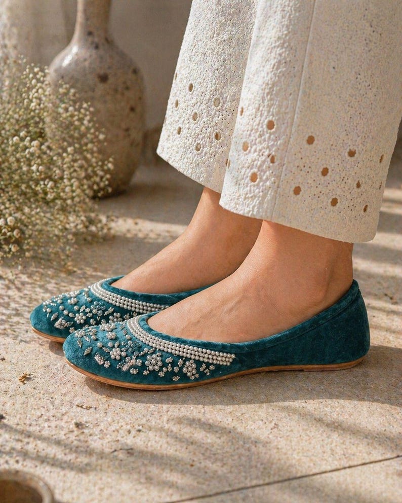 May include: Teal velvet flats adorned with pearls and beads. These shoes feature a rounded toe and a flat sole. The flats are worn with white eyelet trousers, offering a chic and sophisticated appearance.