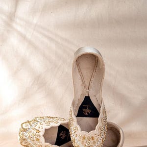May include: A pair of cream-colored slip-on flats featuring intricate gold and silver beadwork, sequins, and embroidery across the upper. The interior features a black insole with a small gold floral line drawing logo printed on the heel area.