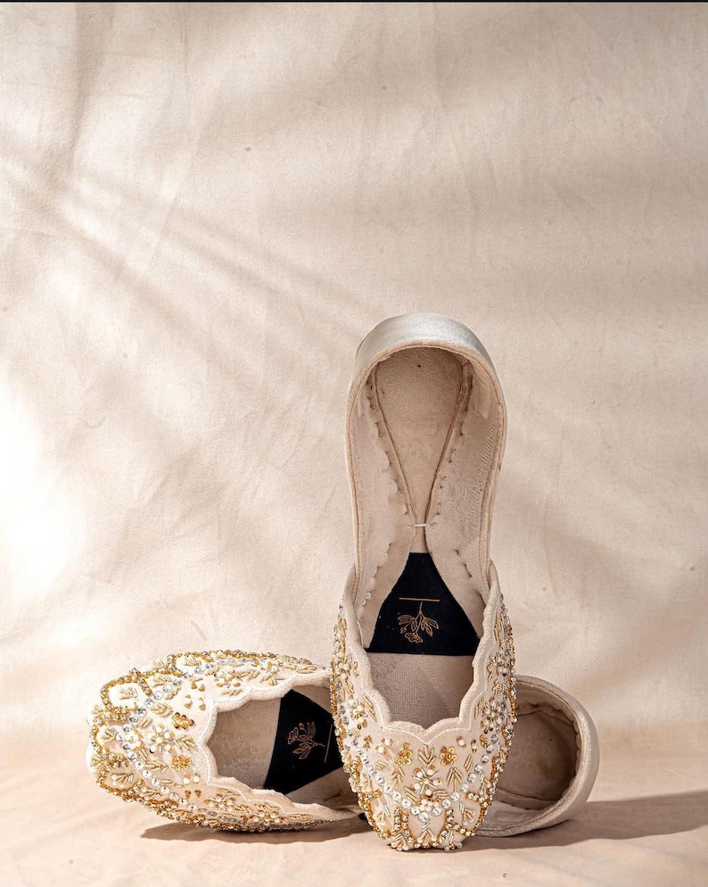 May include: A pair of cream-colored slip-on flats featuring intricate gold and silver beadwork, sequins, and embroidery across the upper. The interior features a black insole with a small gold floral line drawing logo printed on the heel area.