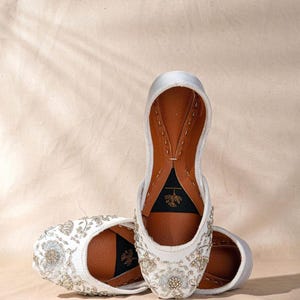 May include: White embroidered slip-on shoes with floral and beaded detailing. The shoes have a leather interior and a soft, off-white exterior. The design features intricate patterns and a classic, elegant style. The shoes are perfect for special occasions.