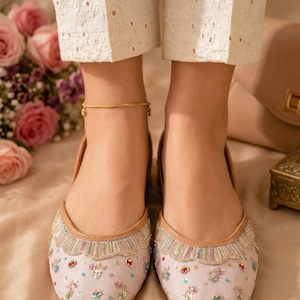 May include: A pair of light lavender flat shoes featuring a scalloped trim along the opening, decorated with multicolored sequins and beads in floral patterns, finished with a tan leather-like binding around the edges of the footwear.