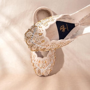 May include: A pair of cream-colored flat shoes featuring a scalloped edge and intricate gold embroidery with clear crystal and pearl embellishments. The interior sole displays a black triangular patch with a gold-colored floral line art illustration.
