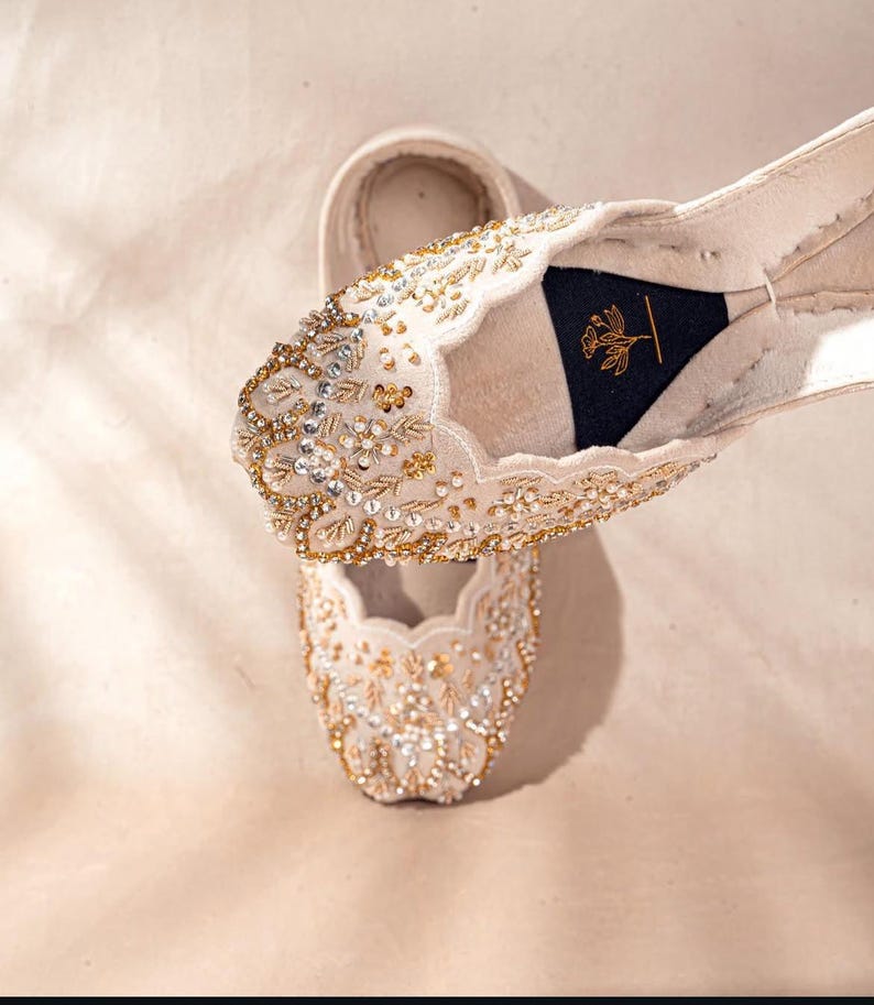 May include: A pair of cream-colored flat shoes featuring a scalloped edge and intricate gold embroidery with clear crystal and pearl embellishments. The interior sole displays a black triangular patch with a gold-colored floral line art illustration.