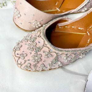 Puede incluir: Primer plano de dos bailarinas rosas con adornos florales. Los zapatos tienen una parte superior de tela rosa claro, adornada con cristales transparentes y brillantes y ribetes plateados. Las suelas son de color marrón claro.