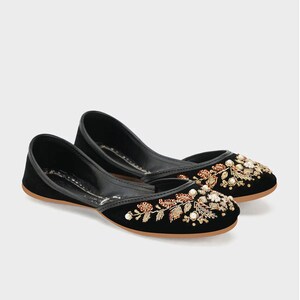 May include: A pair of black velvet flats with intricate floral embroidery in gold, copper, and white beads. The shoes have a rounded toe and a thin, brown sole, showcasing a blend of elegance and craftsmanship.