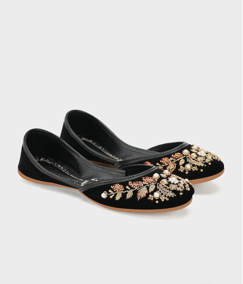 May include: A pair of black velvet flats with intricate floral embroidery in gold, copper, and white beads. The shoes have a rounded toe and a thin, brown sole, showcasing a blend of elegance and craftsmanship.