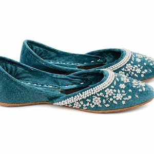 May include: Teal velvet flats with white beaded floral embroidery. The shoes have a rounded toe and a low heel. The upper is decorated with small white beads in a floral pattern.