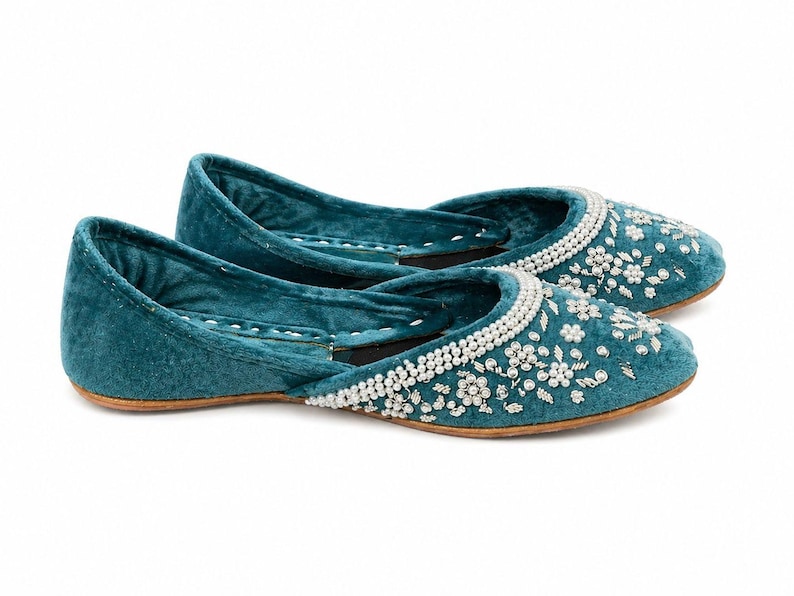 May include: Teal velvet flats with white beaded floral embroidery. The shoes have a rounded toe and a low heel. The upper is decorated with small white beads in a floral pattern.