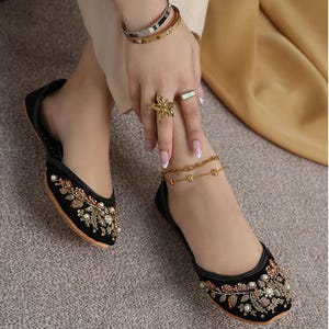 May include: Black velvet flats with gold and pearl embroidery. The flats are adorned with floral and leaf patterns. The image also shows gold bracelets, rings, and an anklet. The background is a neutral color.