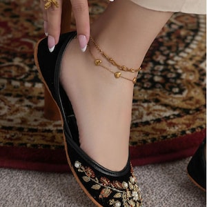May include: Black velvet flats with intricate floral embroidery in gold, red, and white. The shoe features a rounded toe and a thin, contrasting trim. A gold anklet with heart charms and a gold ring with a floral design are also visible.