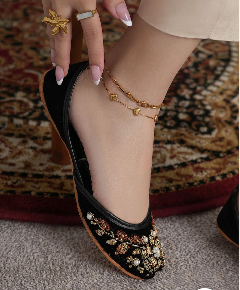 May include: Black velvet flats with intricate floral embroidery in gold, red, and white. The shoe features a rounded toe and a thin, contrasting trim. A gold anklet with heart charms and a gold ring with a floral design are also visible.