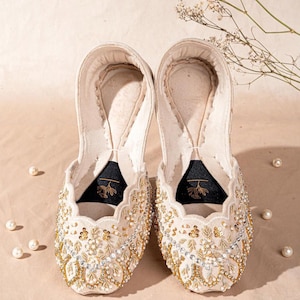 May include: A pair of cream-colored slip-on flats featuring intricate gold embroidery, clear crystal embellishments, and scalloped edges. The interior soles are lined with a dark fabric patch displaying a gold floral graphic and stitching.
