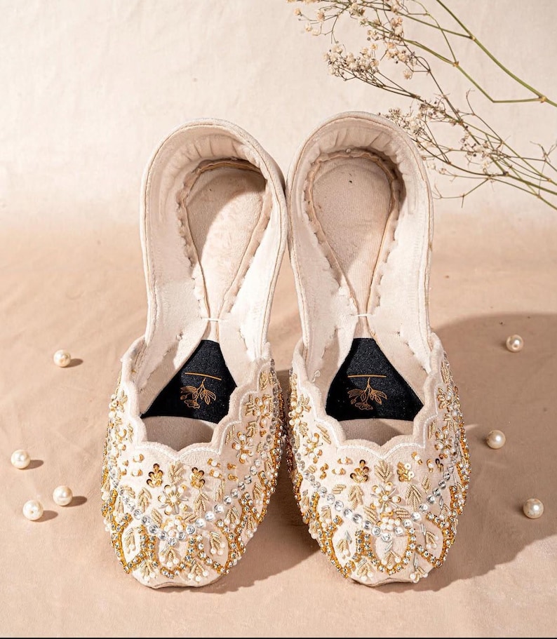 May include: A pair of cream-colored slip-on flats featuring intricate gold embroidery, clear crystal embellishments, and scalloped edges. The interior soles are lined with a dark fabric patch displaying a gold floral graphic and stitching.