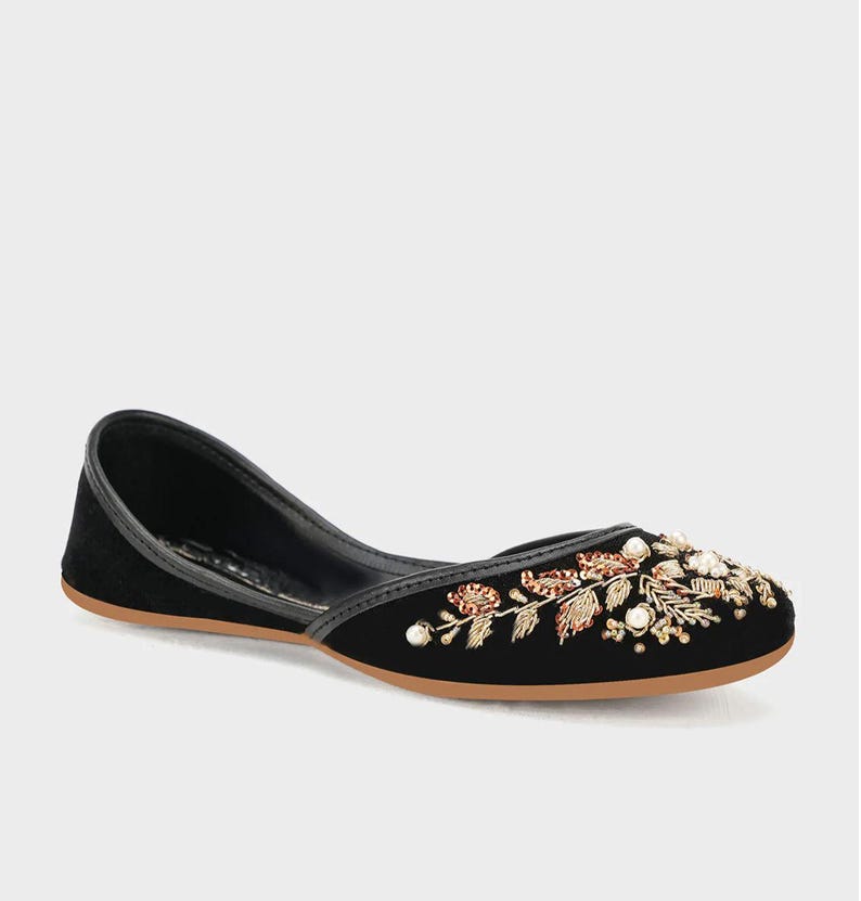 May include: Black velvet flat shoe with floral embroidery. The shoe features a rounded toe and a low heel. The floral design includes gold, red, and white accents. The sole is a light brown color.