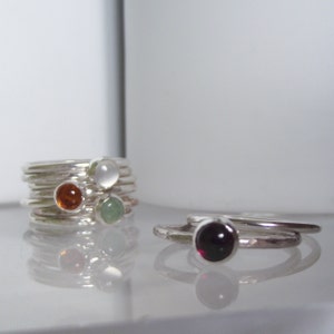 May include: Two silver rings with colourful gemstones. The first ring is a stack of silver bands with a white, green, and orange gemstone. The second ring has a single dark red gemstone.