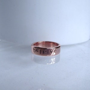 May include: A rose gold band ring with the word "believe" engraved on it.
