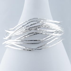 May include: A collection of silver bangles with a textured finish. The bracelets are arranged in a circular formation, with multiple strands creating a layered effect. The jewellery is displayed against a white backdrop.