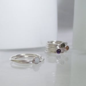 May include: Two silver rings with white stones, one ring is stacked on top of the other. A set of silver rings with different coloured stones is visible in the background.