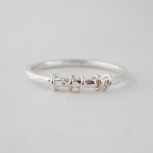 May include: A silver ring with a hammered finish and a gold accent. The ring has a simple design with a band of silver and a small gold circle.