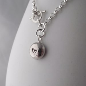 May include: A silver chain necklace with a toggle clasp and a small, round pendant featuring a heart design.