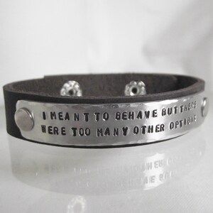 Op de afbeelding: Een bruin leren armband met een zilveren metalen band met de tekst "Ik wilde me gedragen, maar er waren te veel andere opties".